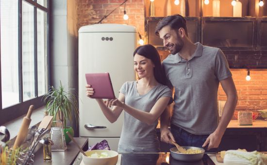 Young beautiful couple in kitchen. Family of two preparing food. Couple using tablet computer while making delicious pasta. Nice loft interior with light bulbs and big window