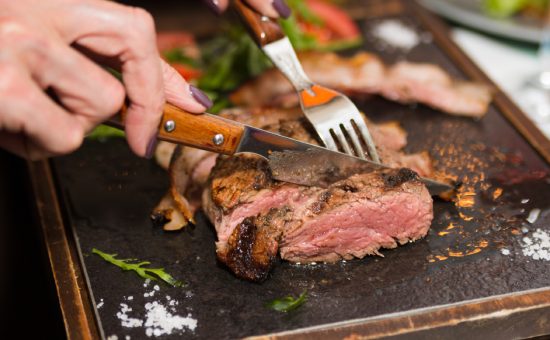 Woman hand holding knife and fork cutting grilled beef steak on stoned plate. Selective focus