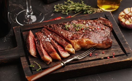 Medium rare sliced grilled striploin beef steak served on wooden board with vintage fork, glass of wine, herbs and spices